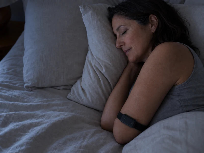 Illustrative nighttime scene of a woman sleeping on her side in a dim bedroom, with a Carbon Mura Band visible on her upper arm